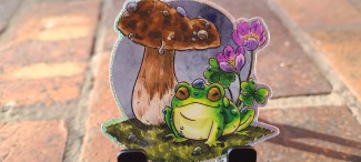 A glittery vinyl sticker of a little green frog happily sitting beneath a mushroom. There are raindrops on the frog, mushroom, and on the sorrel flowers behind.